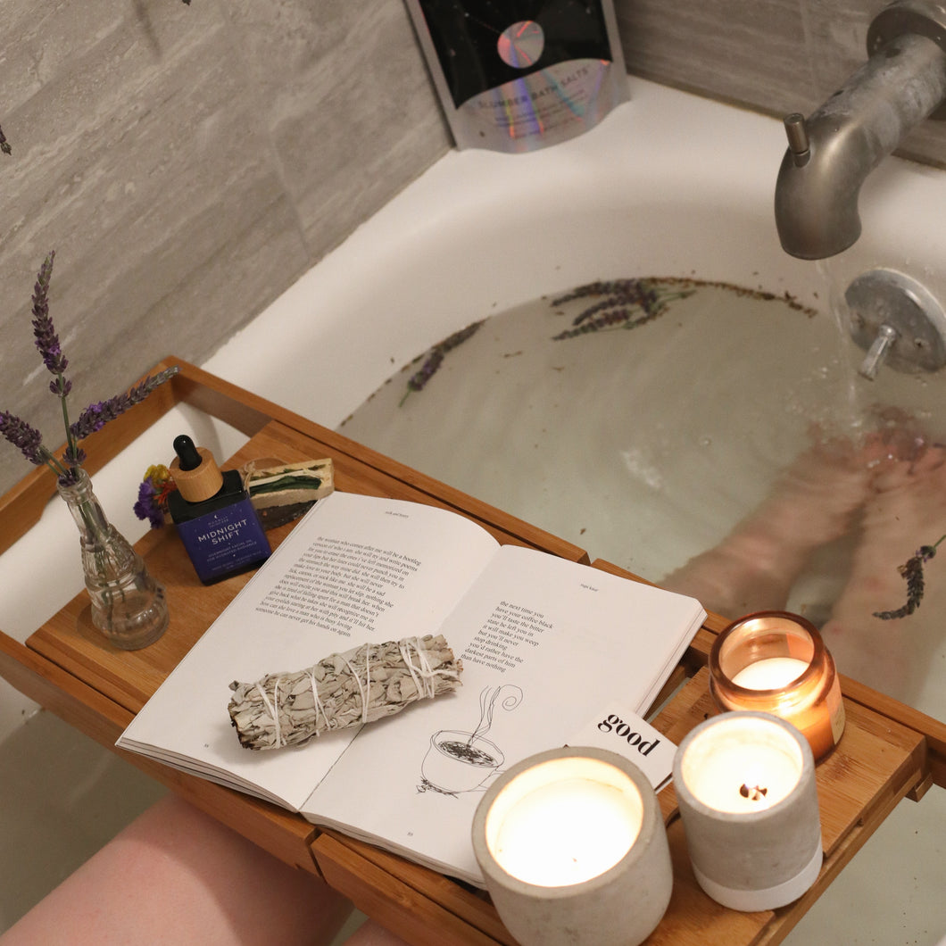 A photo of a tray over a bathtub with candles, a book and a bottle of Midnight Shift Overnight Facial Oil on it