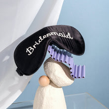 Bridesmaid silk eyemask on stacked rocks and purple paper with blue background and white curtain