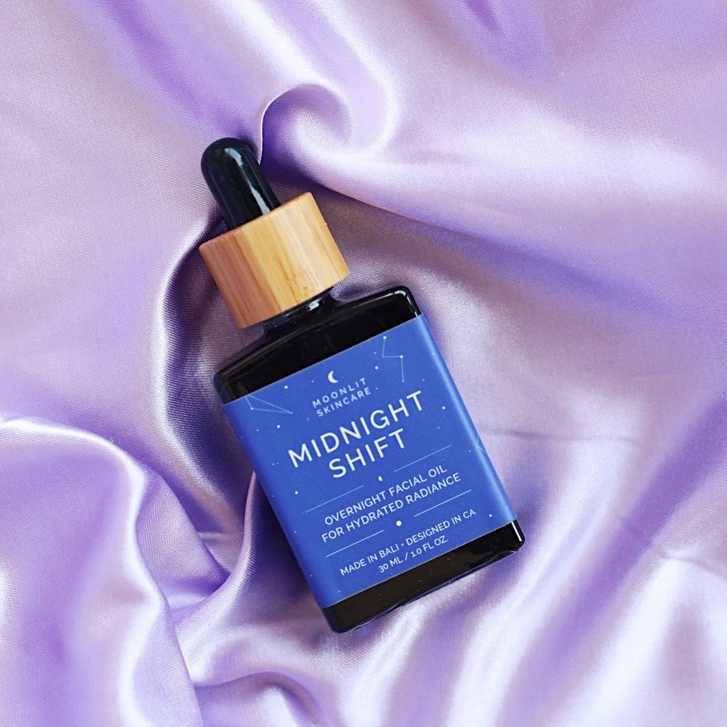 A bottle of Midnight Shift Overnight Facial Oil on a crumpled lilac satin bedsheet
