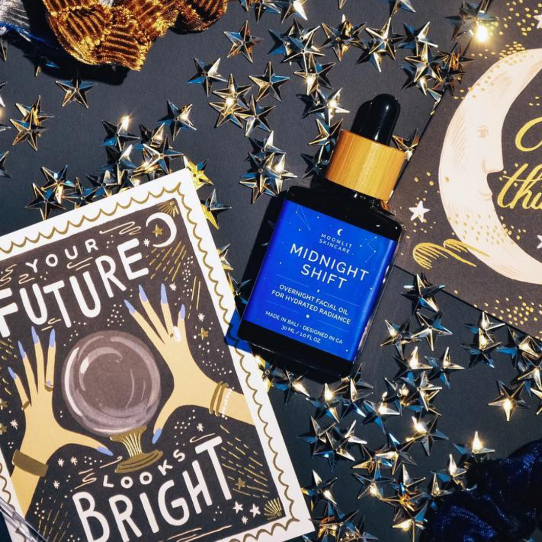 A bottle of Midnight Shift Overnight Facial Oil laid down on a table with tarot cards and start shaped confetti scattered around it