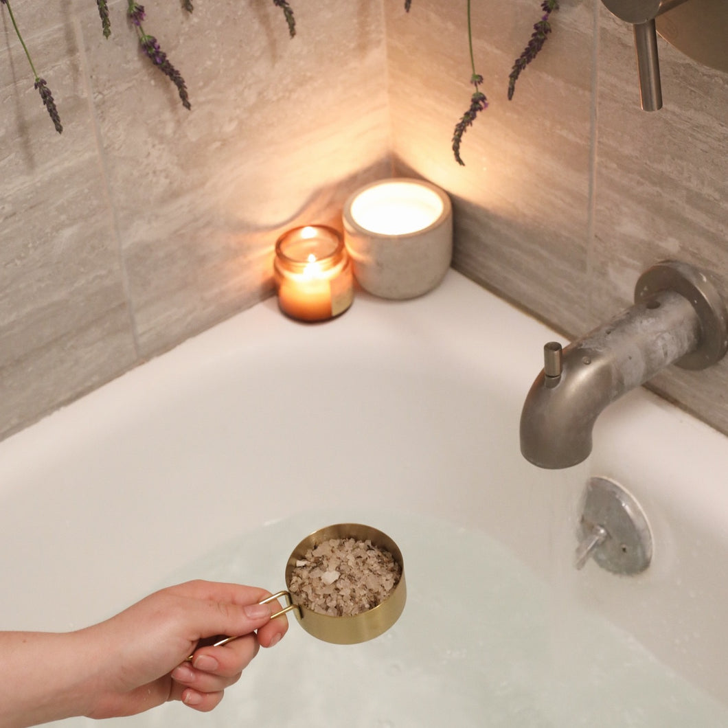 Hand pouring a cup of Slumber Bath Salts into a tub full of water, with two lit up candels on the side of the tub