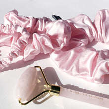 Close up of rose quartz facial roller tool and blush pink silk scrunchie trio set on white background